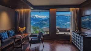 A room with a large window offering a mountain view, a wooden herringbone floor, a sofa with blue cushions, a small table with a vase of flowers, and a television on the wall. - Taj Guras Kutir Resort & Spa, Gangtok (Gangtok)