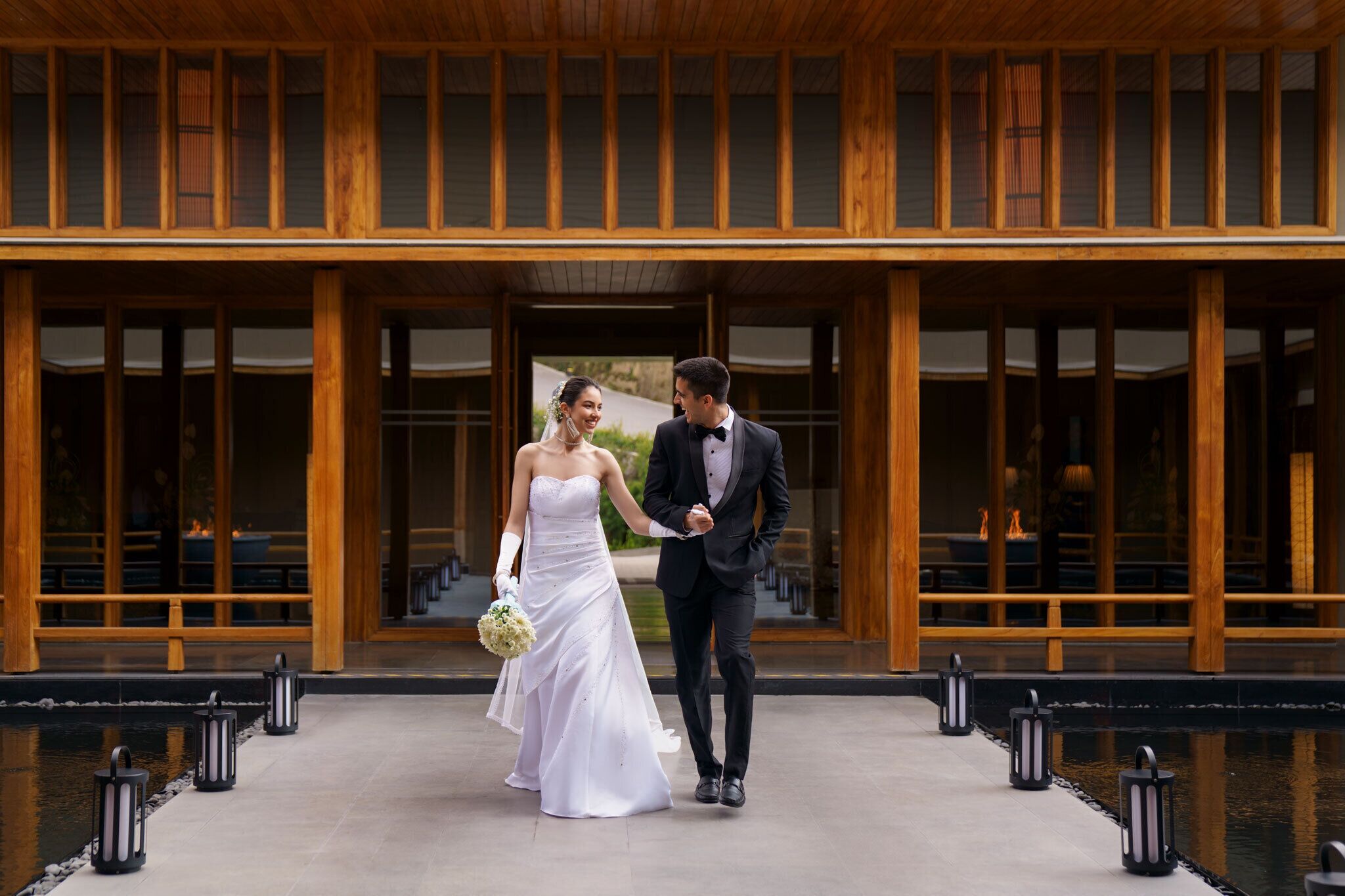 A couple walking hand in hand in front of a building with wooden columns and large windows.