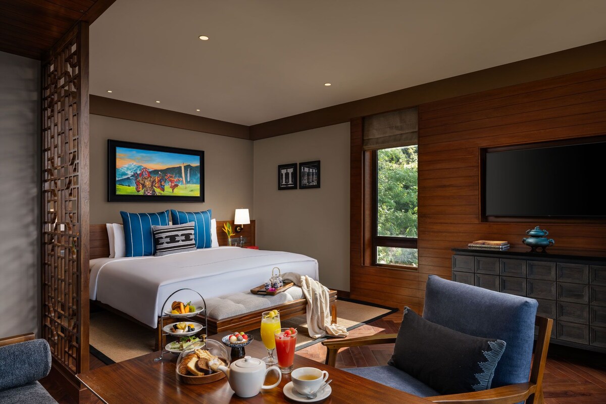 A hotel room with a large bed, a dining table with breakfast, a TV, and a window with a view of greenery.