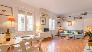 Un salon bien éclairé, avec un canapé, une table à manger dressée pour deux personnes et un téléviseur posé sur un meuble.