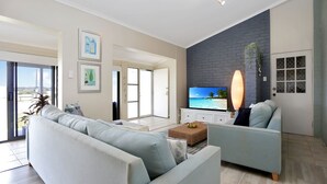A modern living room with a sofa, armchair, and a flat-screen TV mounted on a brick wall. There is a sliding glass door leading to a balcony.