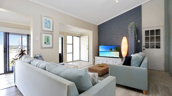 A modern living room with a sofa, armchair, and a flat-screen TV mounted on a brick wall. There is a sliding glass door leading to a balcony.