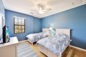 Une chambre avec deux lits simples, un bureau, une chaise, un ventilateur de plafond et une fenêtre avec des stores.