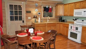 A wooden dining table with red placemats and chairs, a kitchen with wooden cabinets and a white microwave, and a door with a window above it. - Secluded, peaceful Coastal Log Cabin with hot tub. (Townsend)