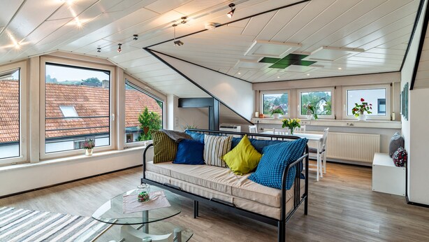 A modern loft-style room with a high ceiling, large windows, a sofa with cushions, a dining table, and a kitchen area. - Holiday Apartment "Andrea" with A/C & Wi-Fi (Bühl)