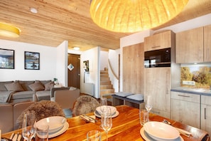 A modern living room with a wooden ceiling, a dining table set for four, and a kitchen with wooden cabinets and a built-in oven.