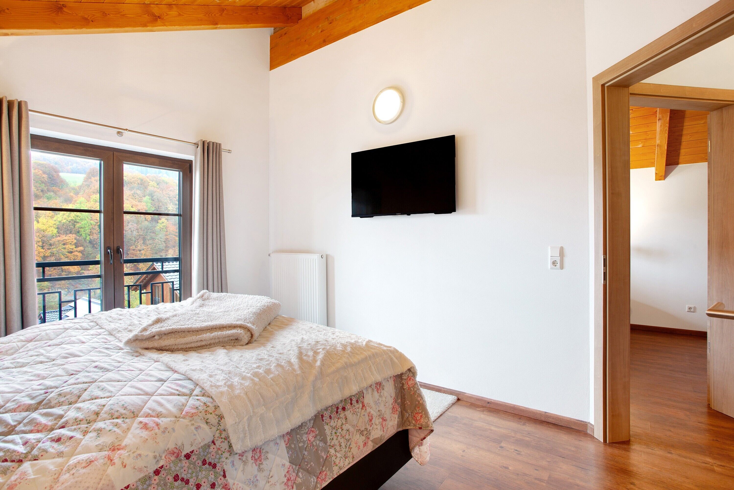 A bedroom with a bed, a television on the wall, a window with curtains, and a view of the outdoors.