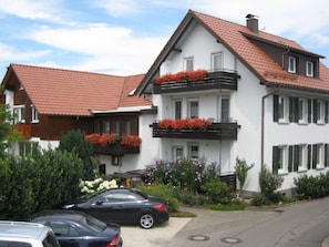 A two-story house with a red-tiled roof, white exterior walls, and a balcony with red flowers.