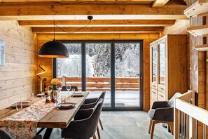 Ein Holzinterieur mit einem Esstisch, Stühlen, einer Pendelleuchte und Blick auf eine verschneite Landschaft durch große Glasüren.