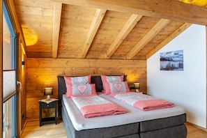 Une pièce chaleureuse avec des poutres apparentes, un lit recouvert d’une couette à carreaux, une table de chevet et une lampe fixée au mur.