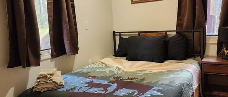 A bedroom with a bed, a nightstand, a window with curtains, a ceiling light, and a framed picture on the wall.