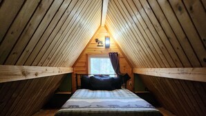 A small, cozy attic bedroom with a wooden sloped ceiling, a single bed with a plaid bedspread, and a window with curtains. - Bear Necessities Cottages (Seward)
