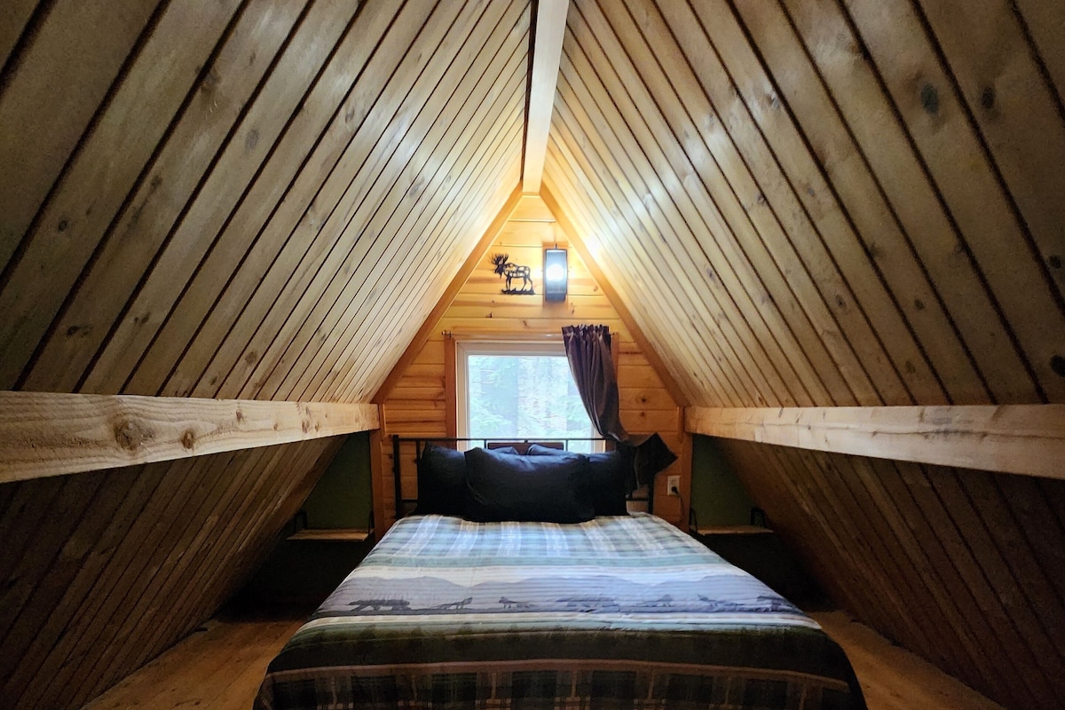 A small, cozy attic bedroom with a wooden sloped ceiling, a single bed with a plaid bedspread, and a window with curtains.
