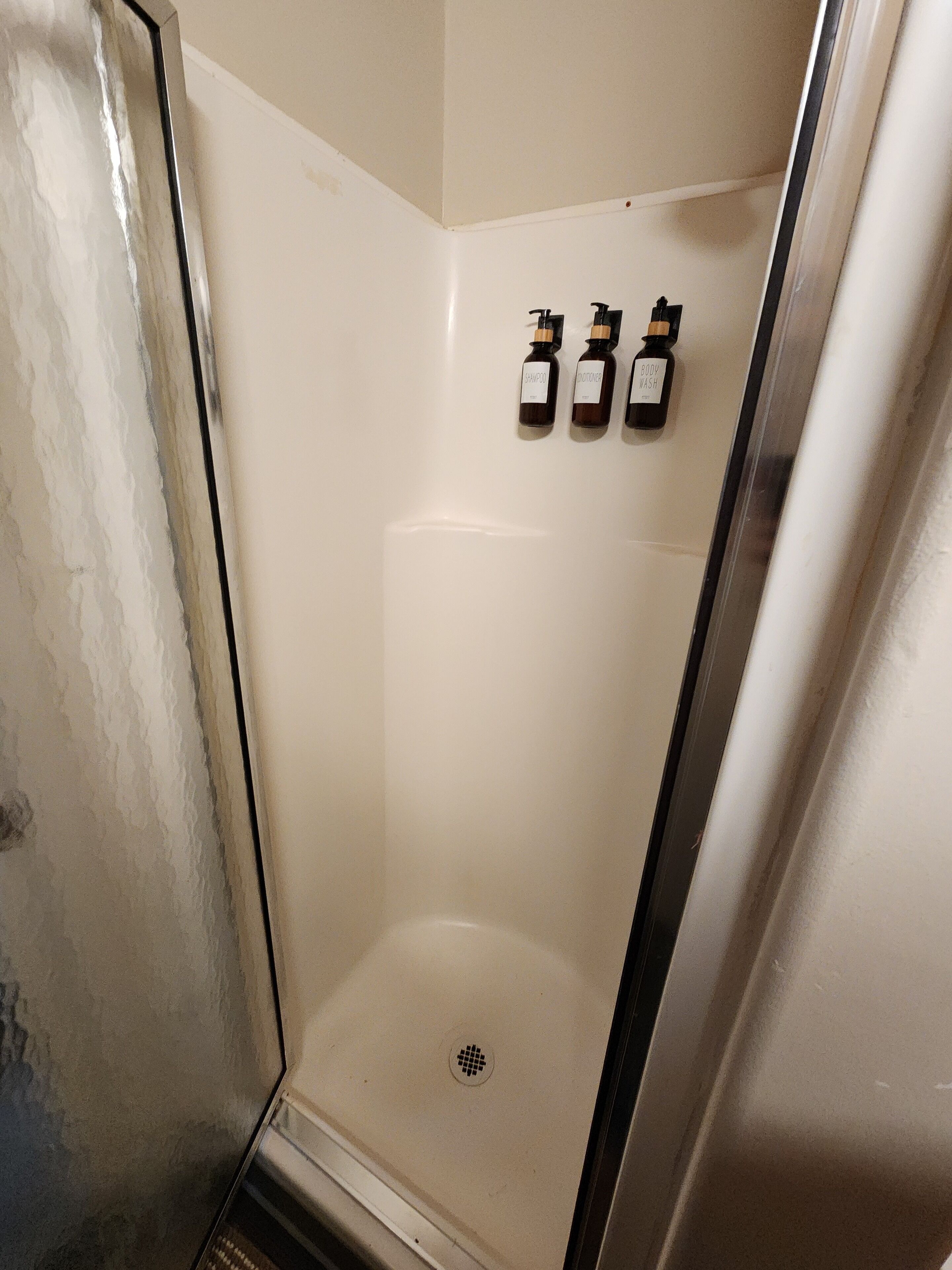 A bathroom with a glass shower enclosure, a white tiled wall, and three bottles of shampoo and conditioner on the wall.