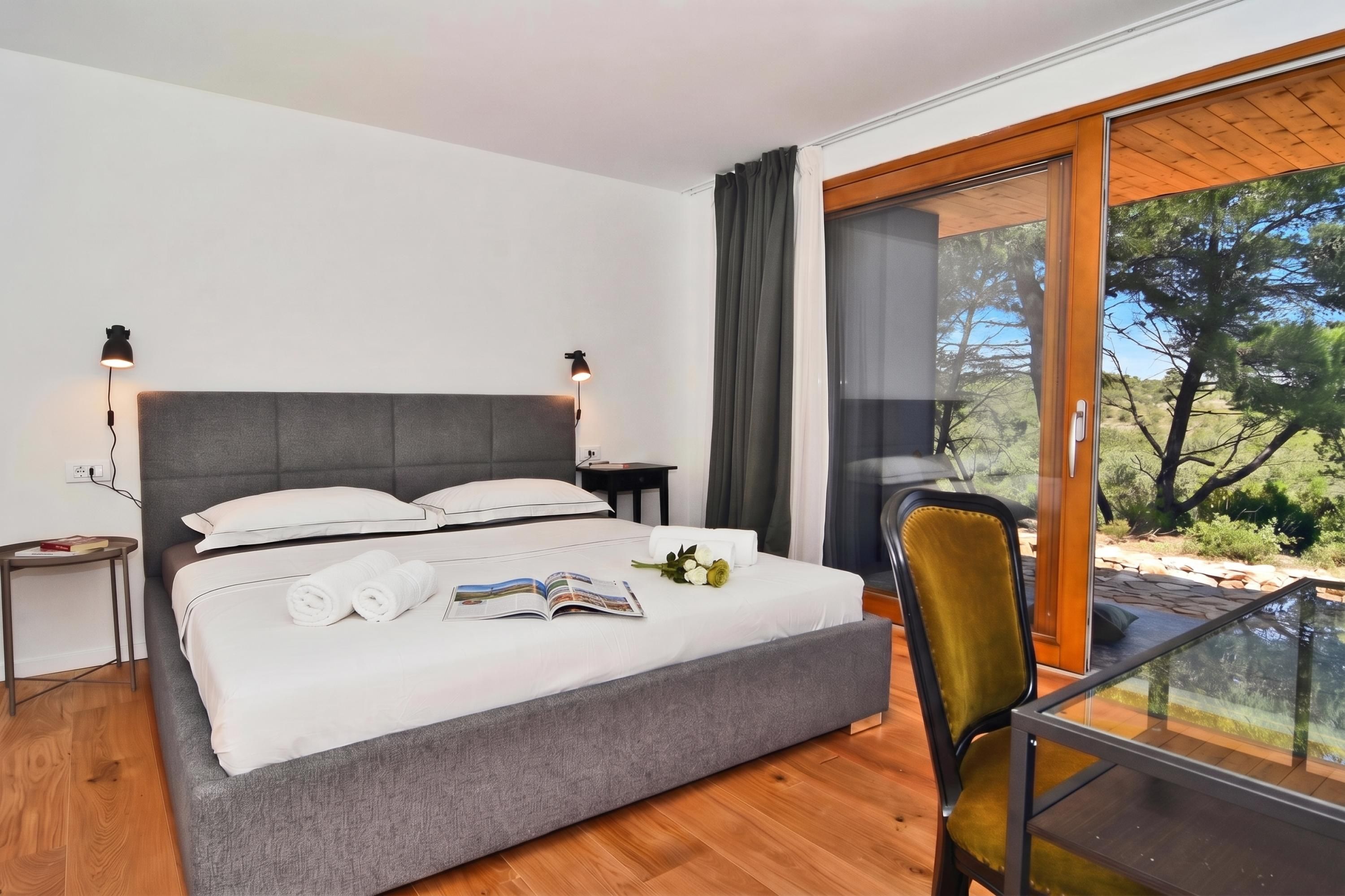 A modern bedroom with a large bed, a glass table, and a view of the outdoors.