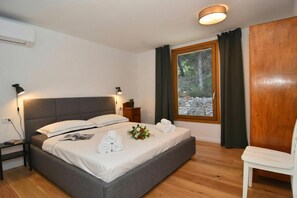 Een opgemaakt bed met kussens, een nachtkastje met een lamp, een raam met gordijnen, en een stoel.