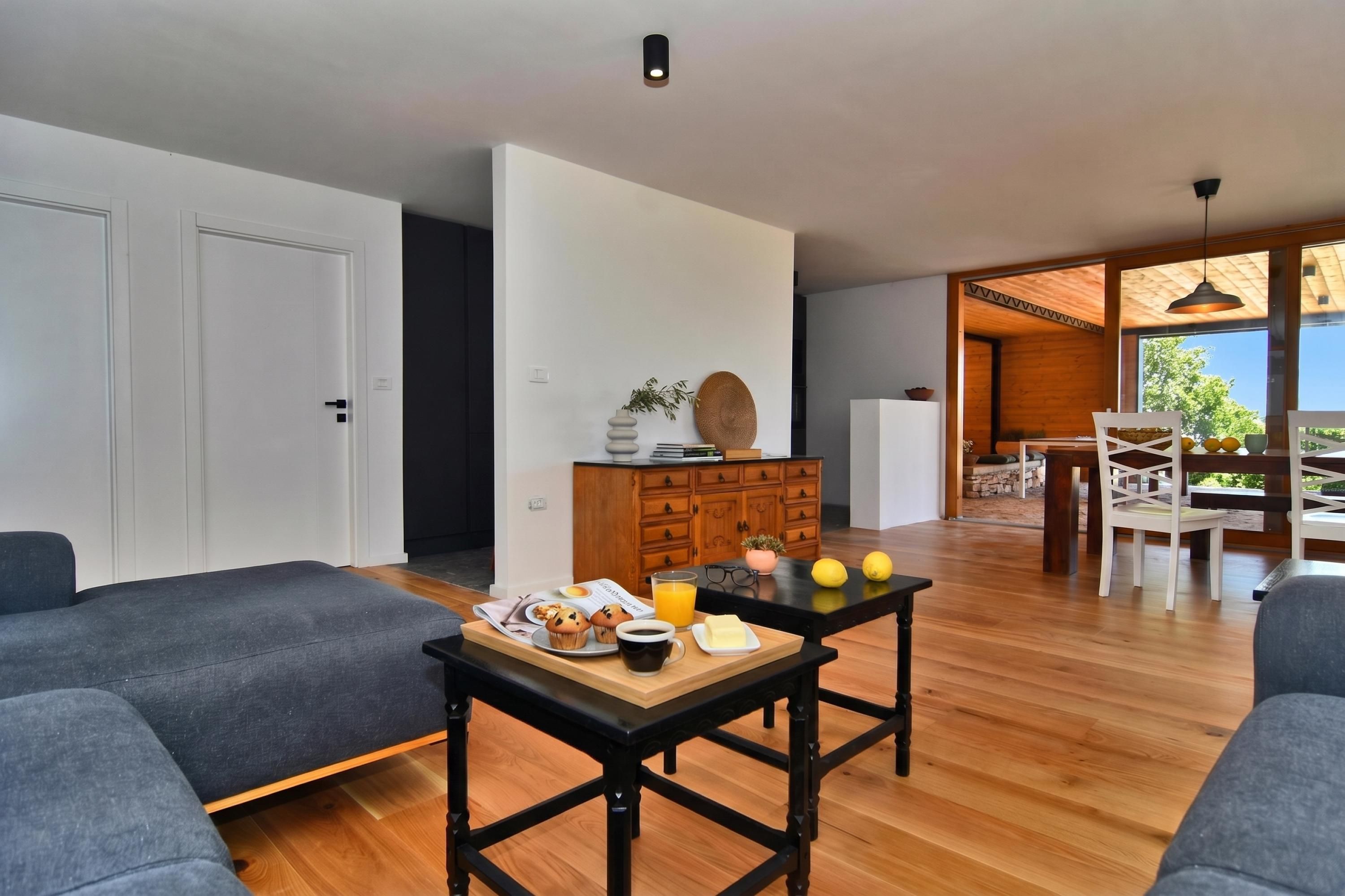 Un salon moderne avec un plancher en bois, des canapés gris, une table à manger où sont disposés des aliments pour le petit-déjeuner, et une vue sur l’extérieur.