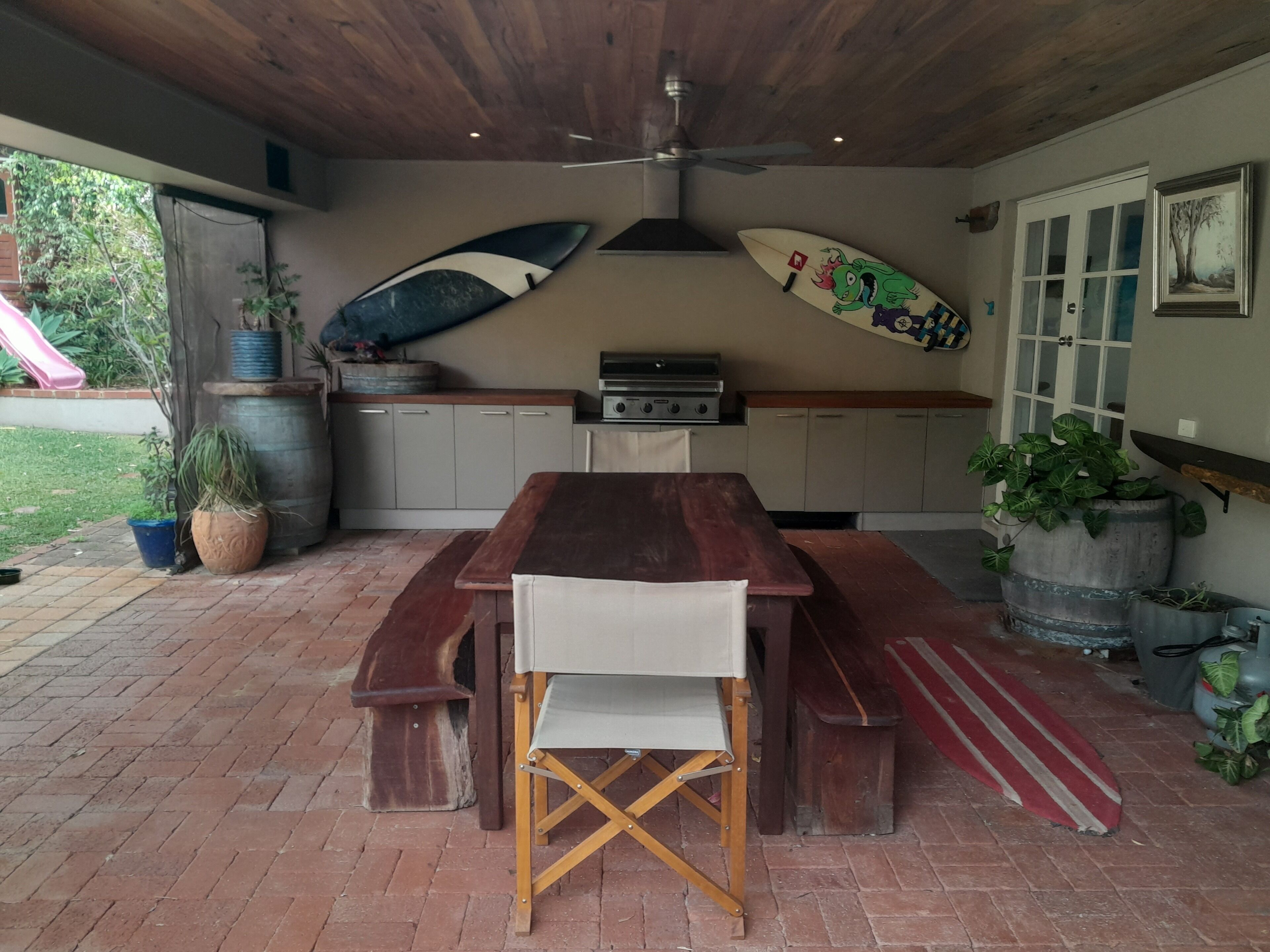 A covered patio with a wooden table and benches, a grill, and surfboards mounted on the wall.