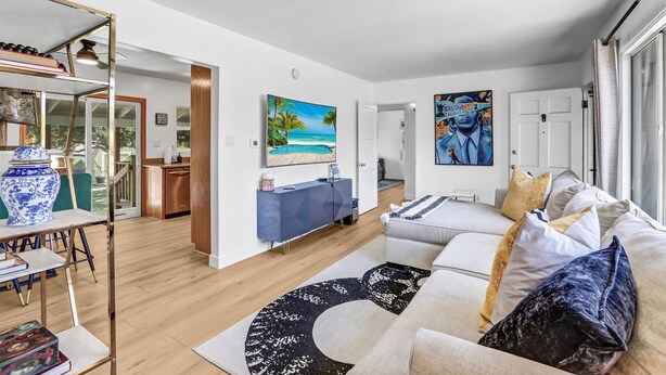 A modern living room with a sofa, a coffee table, and a dining area with a table and chairs. There is a painting on the wall and a door leading to another room.