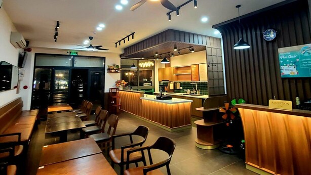 Interior de un restaurante con mesas y sillas de madera, un mostrador de recepción y una cocina visible a través de un separador de vidrio.
