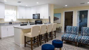 Une cuisine moderne avec des meubles blancs, un îlot central doté de tabourets de bar et un four à micro-ondes. Le salon adjacent est agrémenté de chaises et de poufs recouverts d’un tissu bleu.