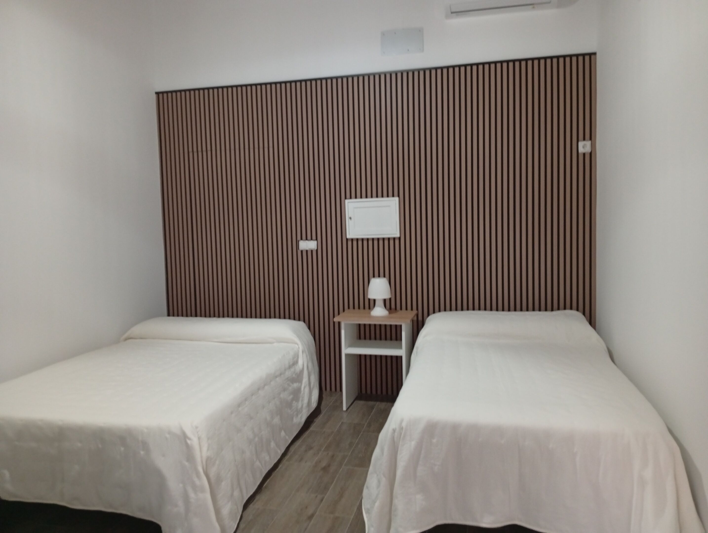 A hotel room with two single beds, a small bedside table with a lamp, and a wall-mounted air conditioning unit.