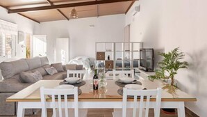 A dining area with a wooden table, white chairs, and a glass-top table. There is a sofa and a television in the background. - Rincón Andaluz, Huerta Alegría (Casariche)