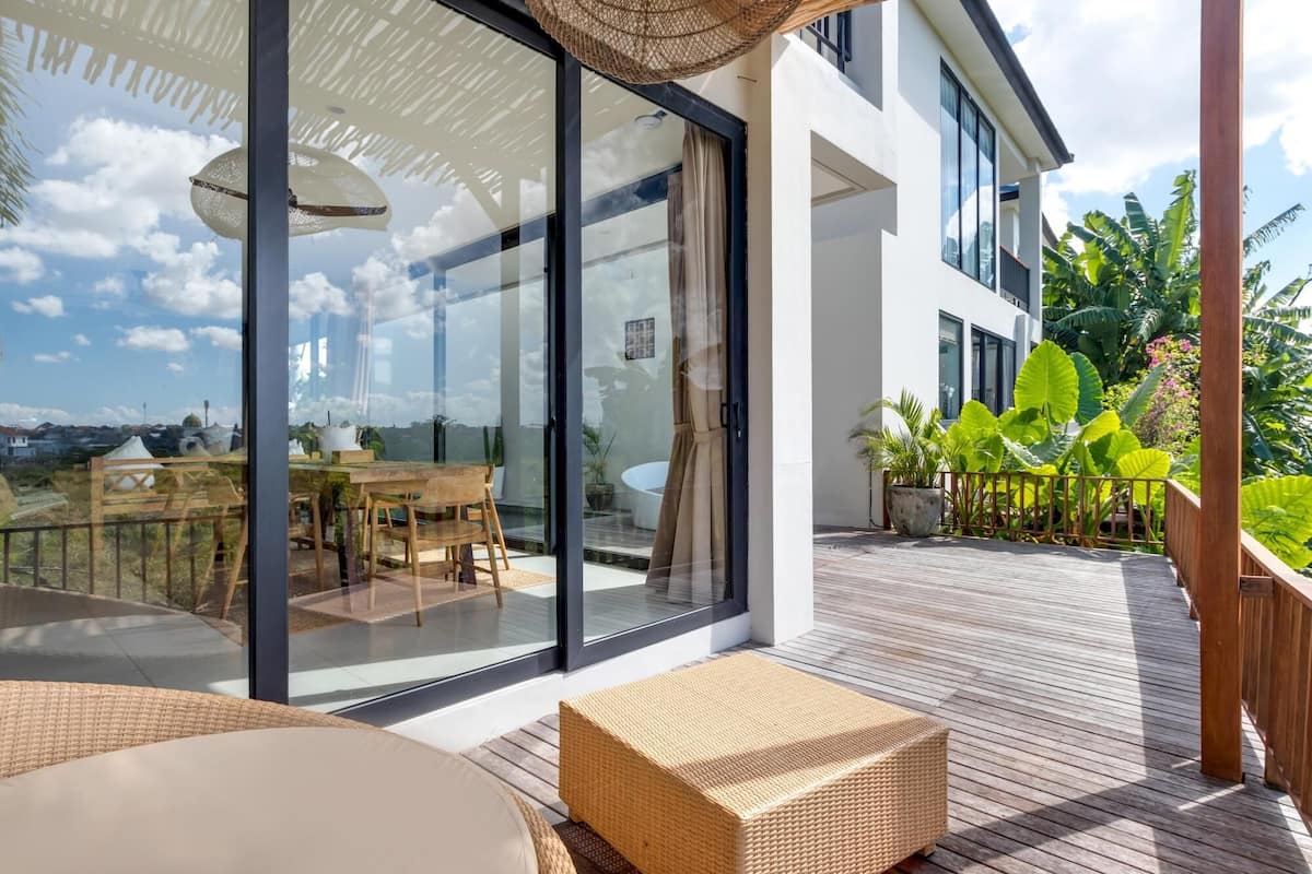 A modern outdoor deck with glass doors leading to a dining area, surrounded by lush greenery and a clear sky.