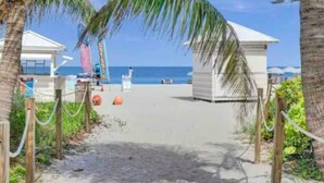 Ein Strandweg führt zu einem Strandabschnitt mit Palmen, einer kleinen Bausubstanz und Blick auf das Meer.