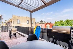 Eine überdachte Terrasse mit Grill, Stühlen und einem Surfbrett.