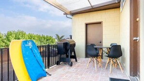 A patio area with a barbecue grill, a table with chairs, and a surfboard leaning against the wall.