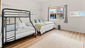 A room with a bunk bed, a single bed, a window with curtains, a ceiling fan, and a view of a building outside.