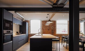 Una cocina moderna con armarios oscuros, una isla central y un comedor con pared de ladrillo y ventanas amplias.