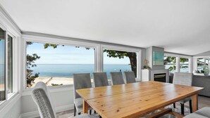 A dining area with a wooden table and grey upholstered chairs, large windows with a view of the sea, and a grey sofa in the background. - Crystal Beach Waterfront Cottage - Sunset Views! (Fort Erie)