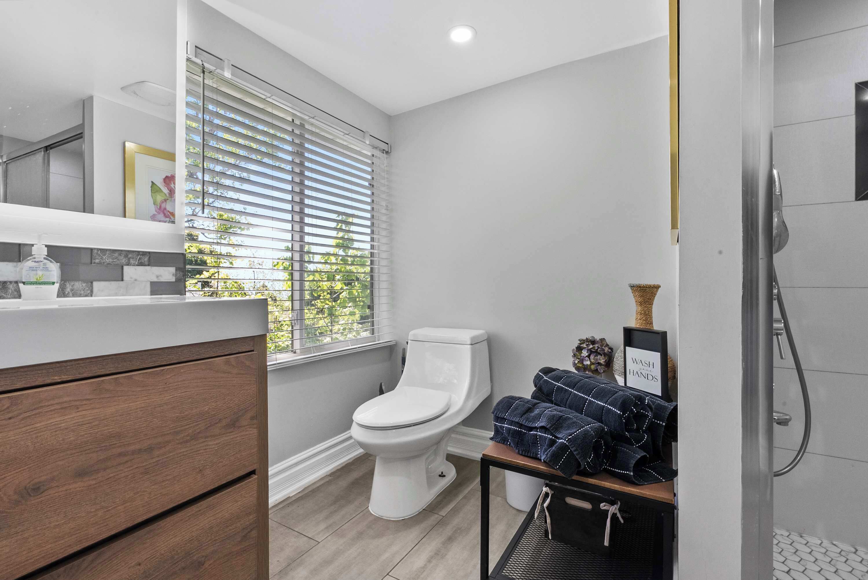 Ein modernes Badezimmer mit Toilette, einem Waschbecken mit Holzschrank, einem Fenster mit Jalousien und einer Duschkabine.