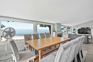 Un salon moderne avec une table à manger en bois, des chaises à revêtement gris, une grande fenêtre donnant sur la mer, un ventilateur sur pied et un téléviseur à écran plat.