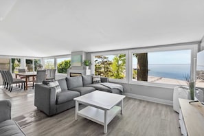 Ein modernes Wohnzimmer mit einem grauen Sofa, einem weißen Couchtisch und großen Fenstern mit Meerblick.
