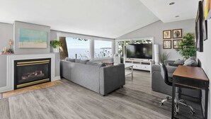 A modern living room with a fireplace, grey sofas, a TV, and large windows. - Crystal Beach Waterfront Cottage - Sunset Views! (Fort Erie)
