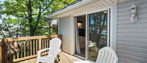 A deck with two white Adirondack chairs, a wooden railing, and a sliding glass door leading to an interior space.
