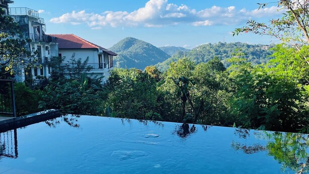 Une piscine agrémentée d’un panneau d’avertissement, entourée de carreaux de pierre et de verdure, avec vue sur des montagnes et un bâtiment en arrière-plan.