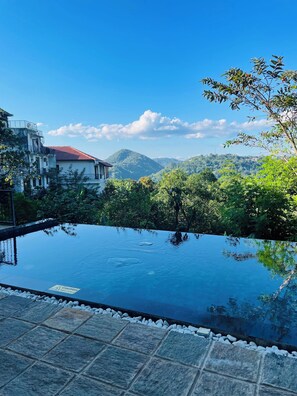 A swimming pool with a caution sign, surrounded by stone tiles and greenery, with a view of mountains and a building in the background. - Amandara Hills Kandy (Kandy)