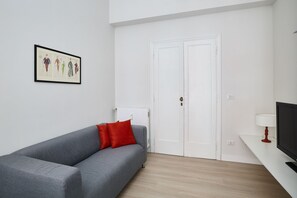 Ett modernt vardagsrum med en grå soffa, röda kuddar, ett inramat konstverk, en vit dörr och en tv på ett vitt tv-bord.