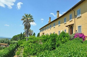 Um edifício grande e amarelo com várias janelas e uma chaminé imponente, rodeado por vegetação exuberante e uma palmeira.
