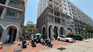En byggnad med flera våningar och balkonger, ett parkeringsområde med motorcyklar och en vit bil som står parkerad på gatan.