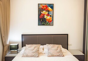 Ein ordentlich bezogenes Bett mit zwei Zierkissen, ein Nachttisch mit Lampe und einer Schachtel, sowie ein Blumenbild an der Wand.