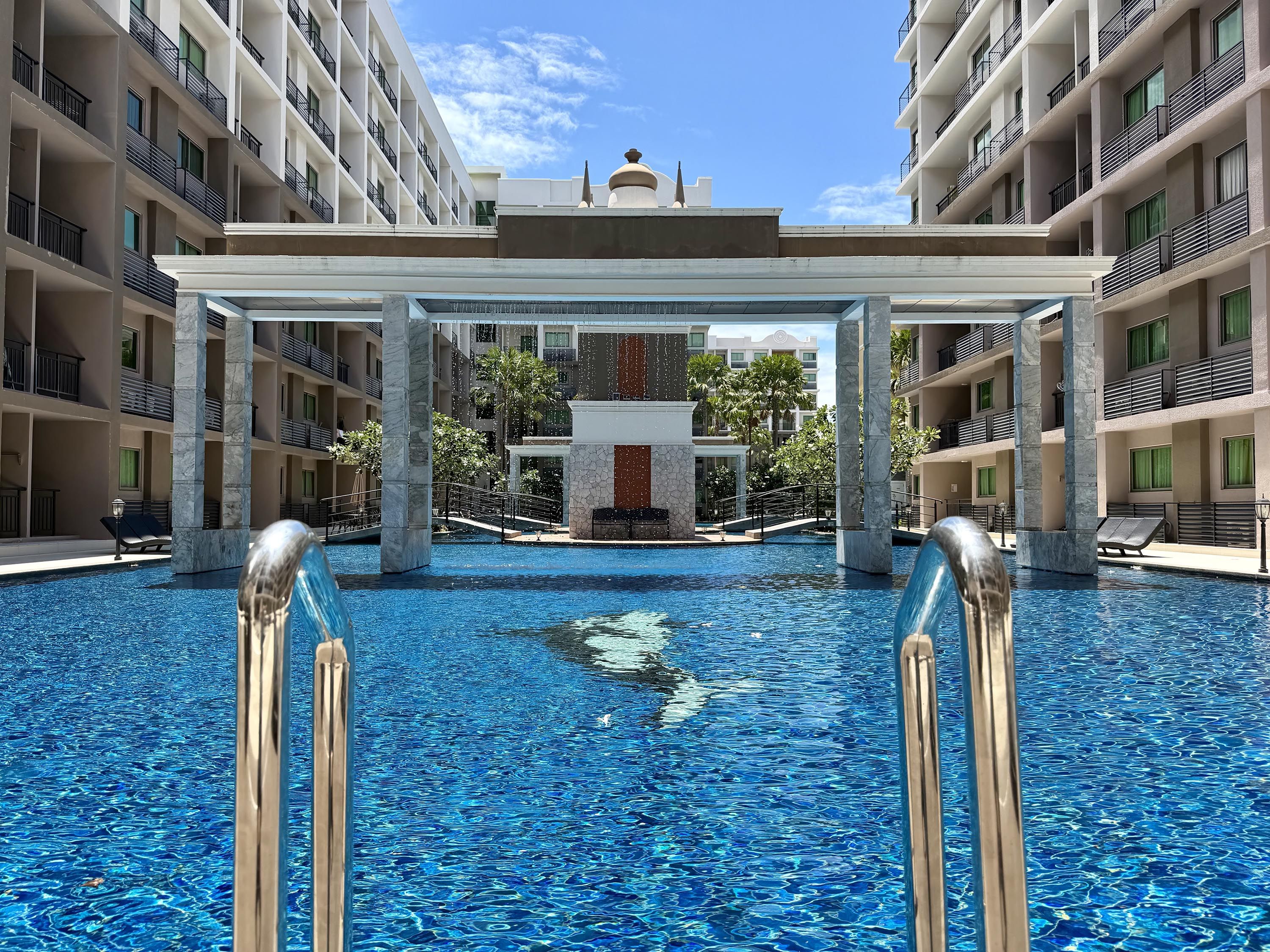 A hotel pool area with a clear view of the pool, a covered walkway, and a central fountain.