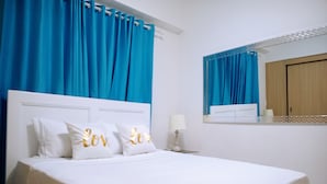 Una cama bien hecha con sábanas blancas y dos almohadas con el texto 'Love' bordado. Se ve una mesita de noche con una lámpara, un espejo sobre la cama y una puerta de madera al fondo.