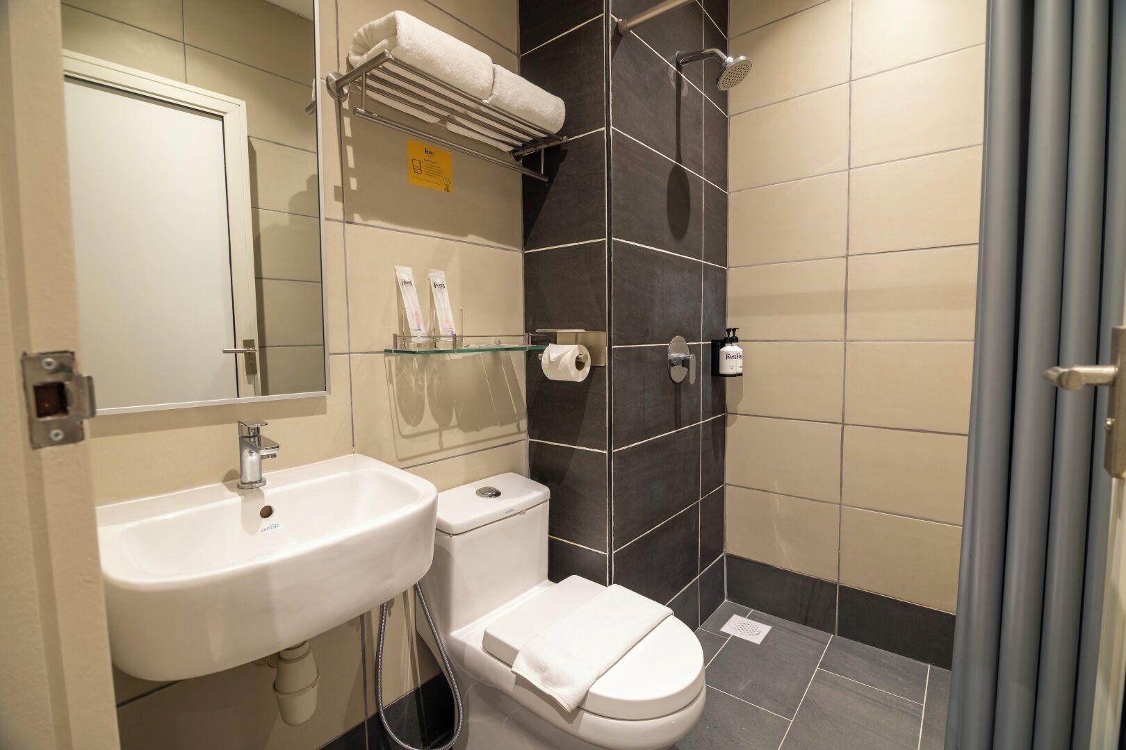 Ein modernes Badezimmer mit Waschbecken, Toilette und Dusche. Eine Glasablage mit Toilettenartikeln und ein Handtuchhalter befinden sich über dem Waschbecken.
