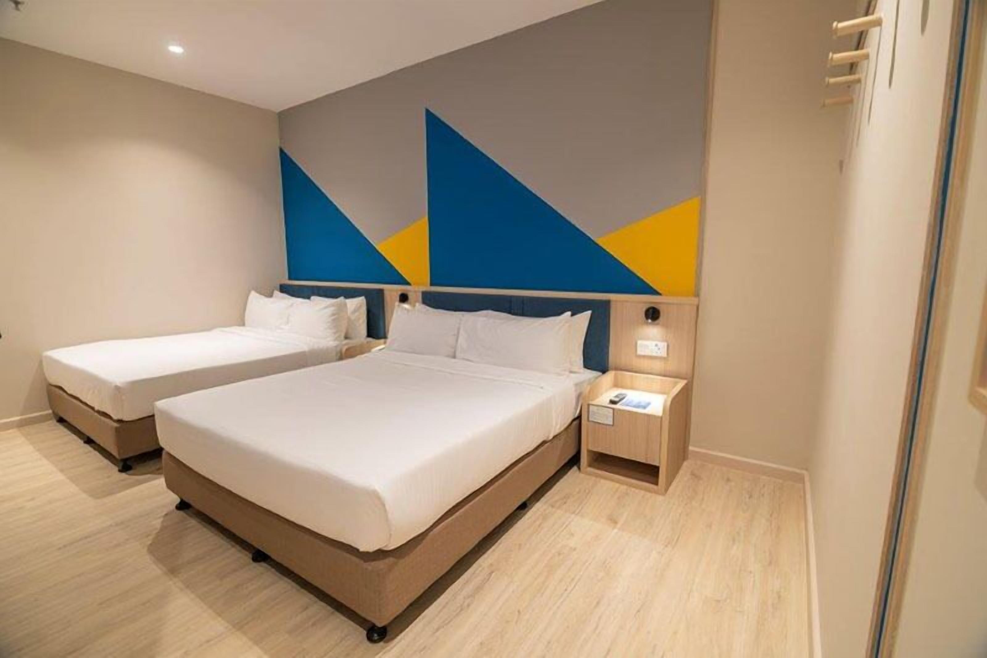 A hotel room with two beds, a bedside table with a lamp, and a wall with geometric patterns.