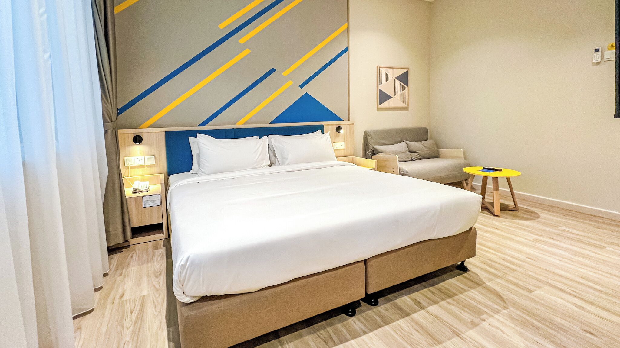 A modern hotel room with a large bed, a bedside table with a lamp, a small sofa, and a side table with a phone.